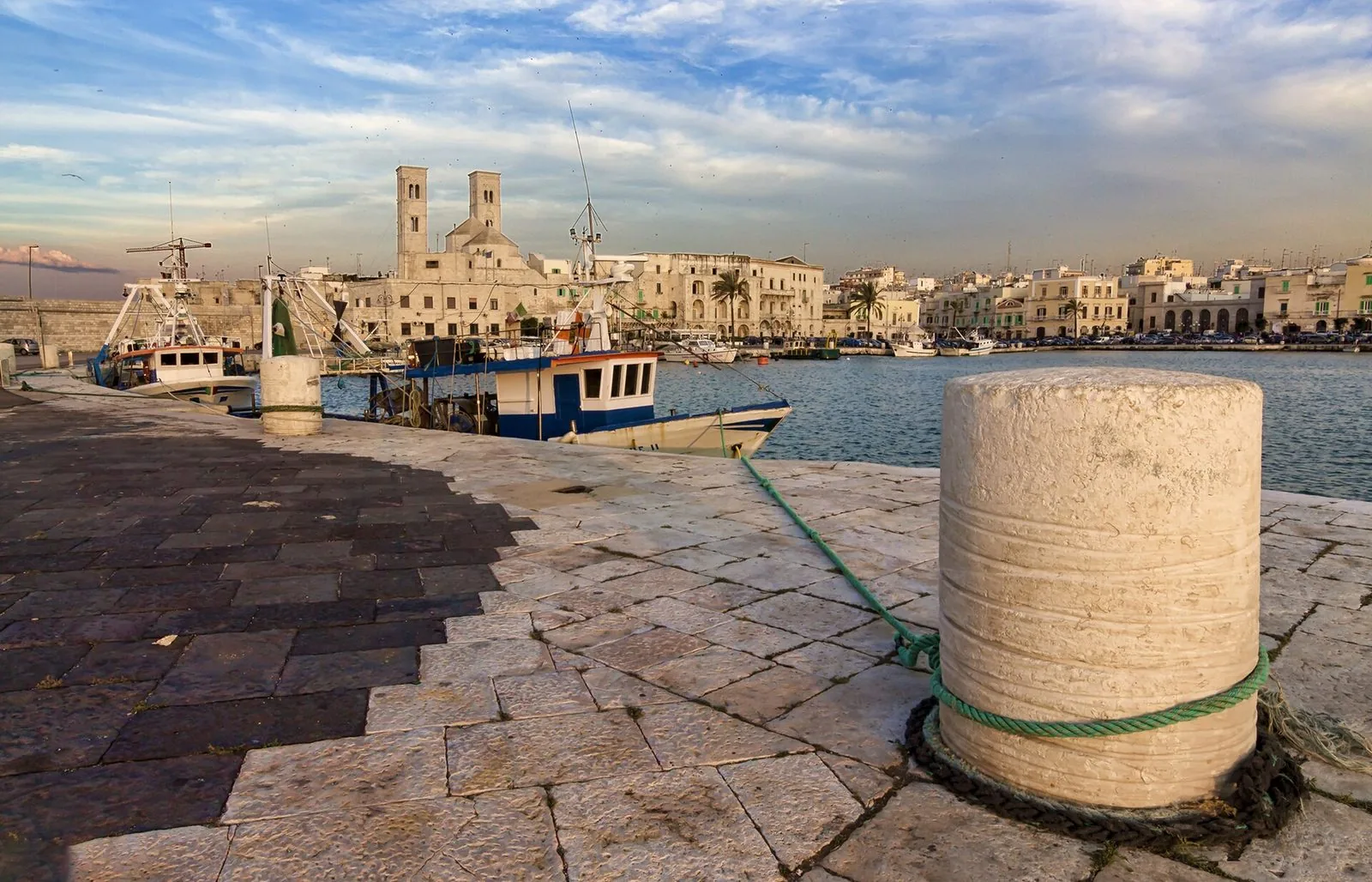 Il porto di Molfetta con i pescherecci ormeggiati e, sullo sfondo, le due torri del Duomo vecchio di San Corrado; in primo piano una bitta in pietra sulla banchina
