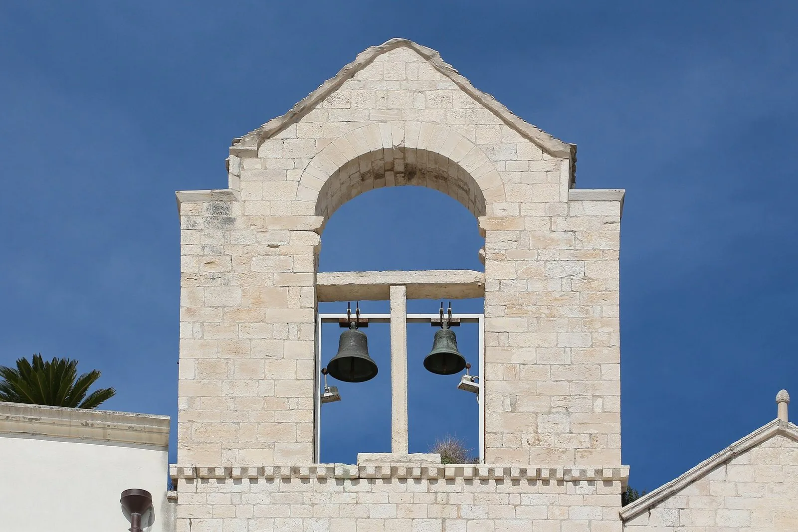 Il campanile a vela della chiesa di Ognissanti a Trani: un'arcata in conci di pietra chiara con due campane di bronzo, sotto un tetto a capanna, contro un cielo blu intenso