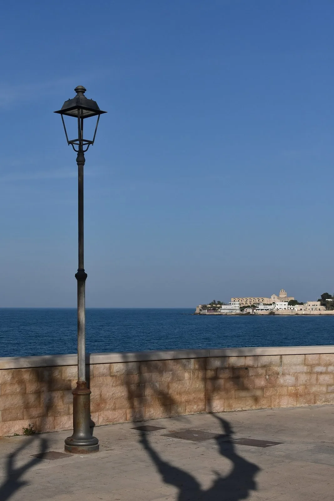 Il lungomare di Trani con un lampione in ferro e il muretto in pietra; sul mare blu, in lontananza, la penisola di Colonna con il complesso dell'abbazia di Santa Maria