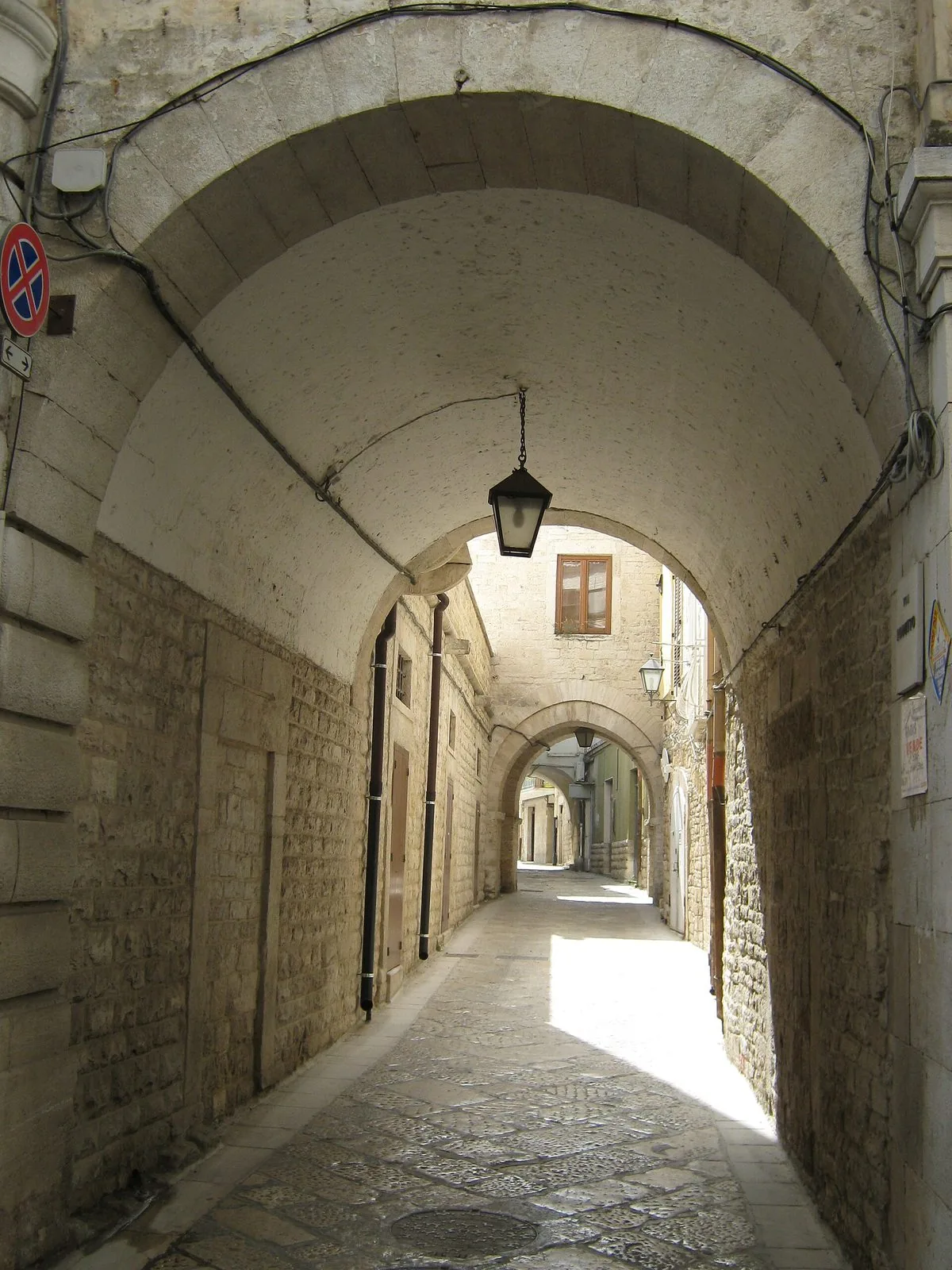 Un vicolo del centro storico di Trani passa sotto un arco di pietra chiara con una lanterna appesa, e prosegue fra case in conci di calcare