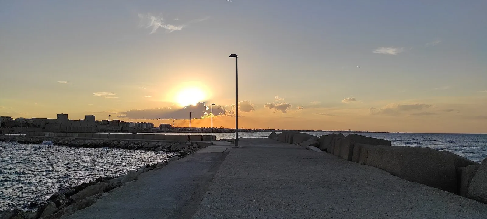 Il molo di Trani deserto al tramonto, con il sole basso tra le nuvole, il mare calmo e il profilo del Castello Svevo sullo sfondo